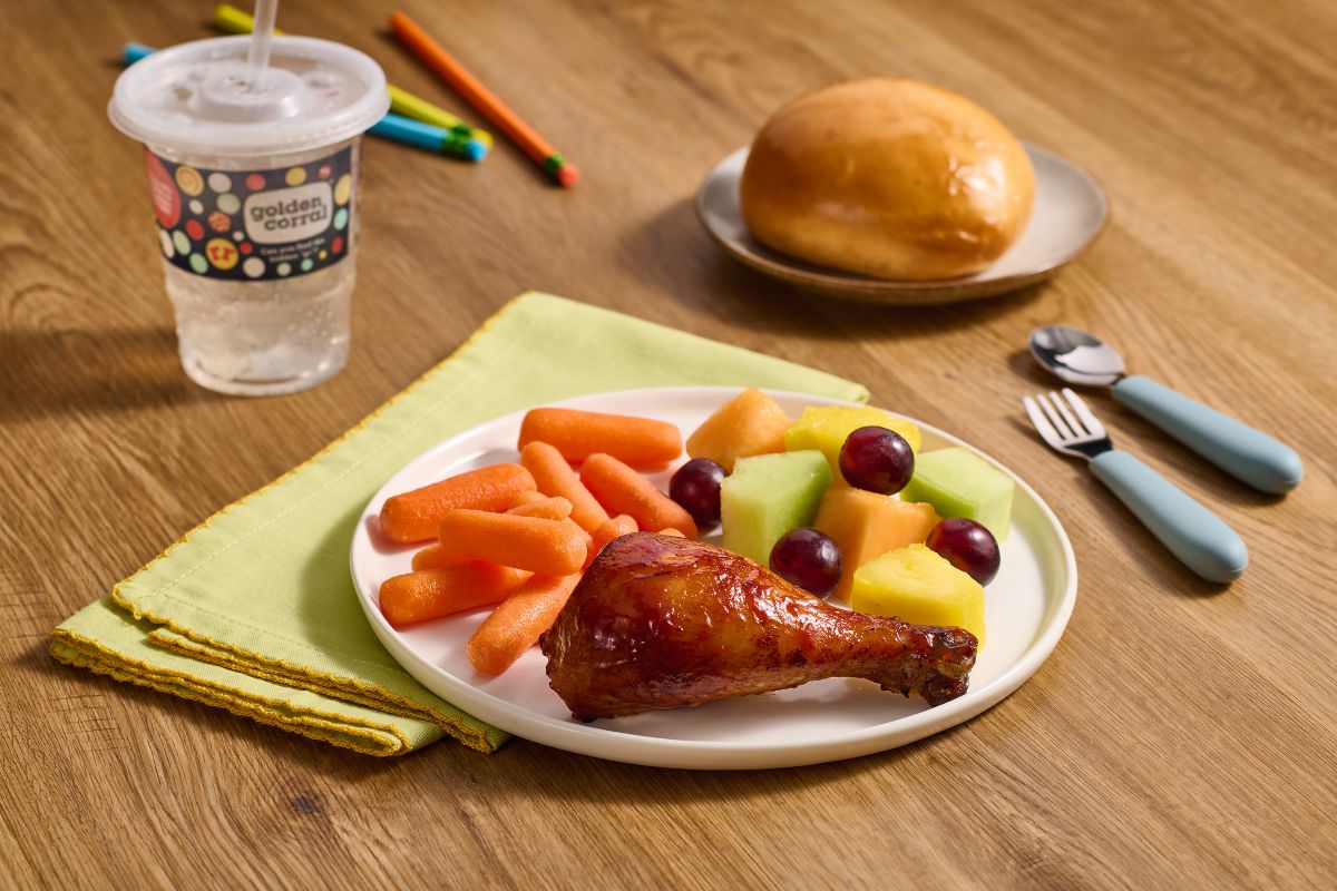 A plate with a roasted chicken drumstick, baby carrots, and fruit salad sits on a green napkin next to a Golden Corral kids deal cup, bread, and utensils.