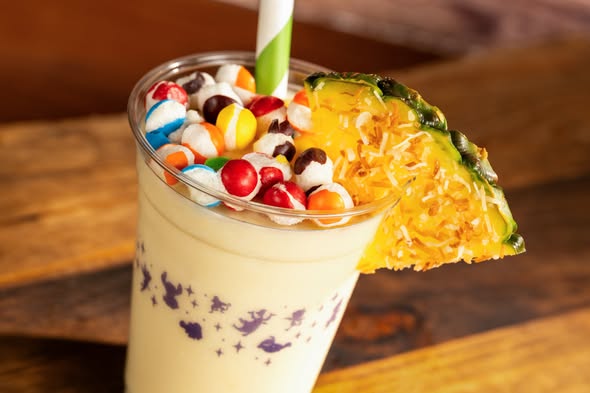 A close-up of a person holding a white cup filled with purple ube soft-serve ice cream, topped with vibrant diced mango, red fruit chunks, and shredded coconut.