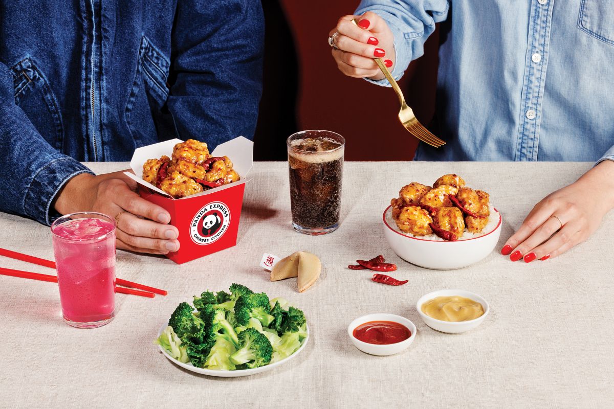 Two people sitting at a table sharing a Panda Express meal featuring Hot Orange Chicken with dried red chili peppers. The table includes a red takeout box and a white bowl filled with orange chicken, a plate of steamed broccoli, a fortune cookie, dipping sauces, a glass of dark soda, and a glass of pink beverage.