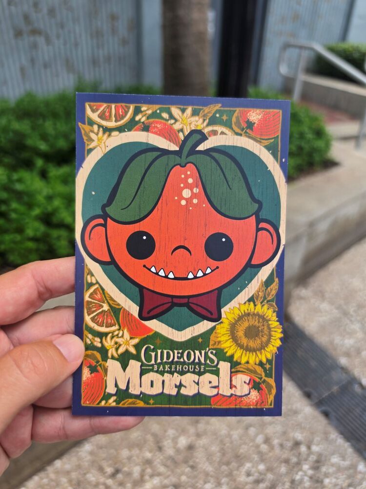 A hand holding the front of a Gideon's Bakehouse July menu card featuring an illustration of an orange-headed character with green hair and a bow tie, framed by sunflowers and orange slices, with the words "Gideon's Bakehouse Morsels" at the bottom.