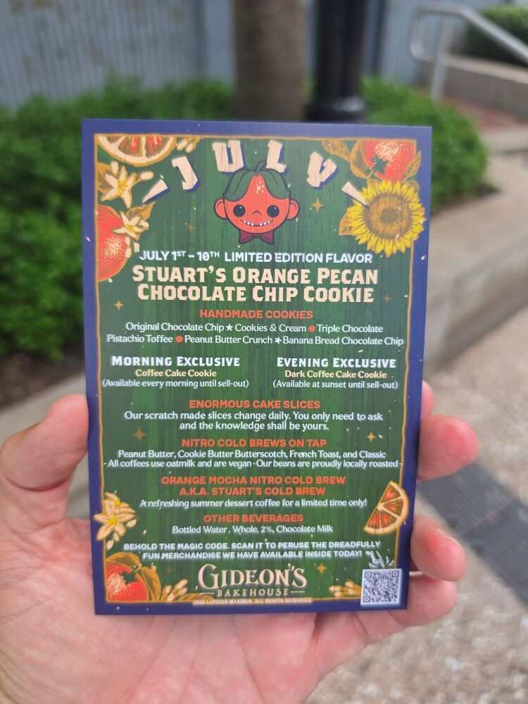 A hand holding the back of a Gideon's Bakehouse July menu card detailing limited-edition items like Stuart's Orange Pecan Chocolate Chip Cookie, handmade cookies, morning and evening exclusives, cold brews, and a QR code at the bottom.