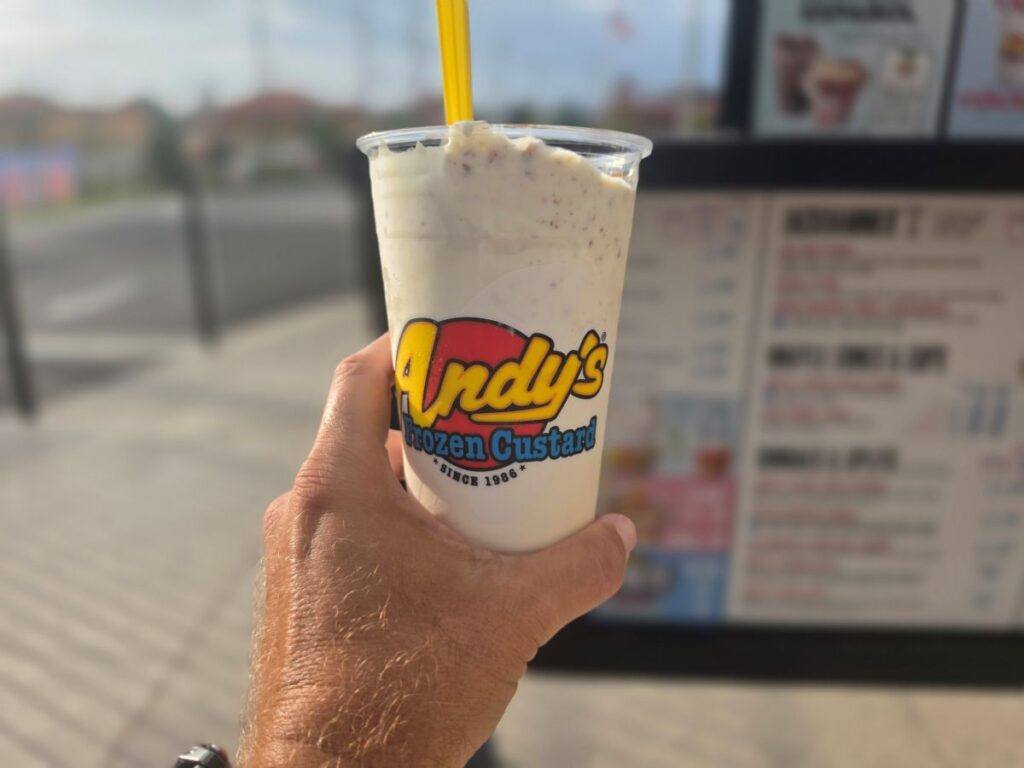 A hand holding a clear cup filled with a Key Lime Pie Concrete treat from Andy's Frozen Custard with a yellow spoon inserted.