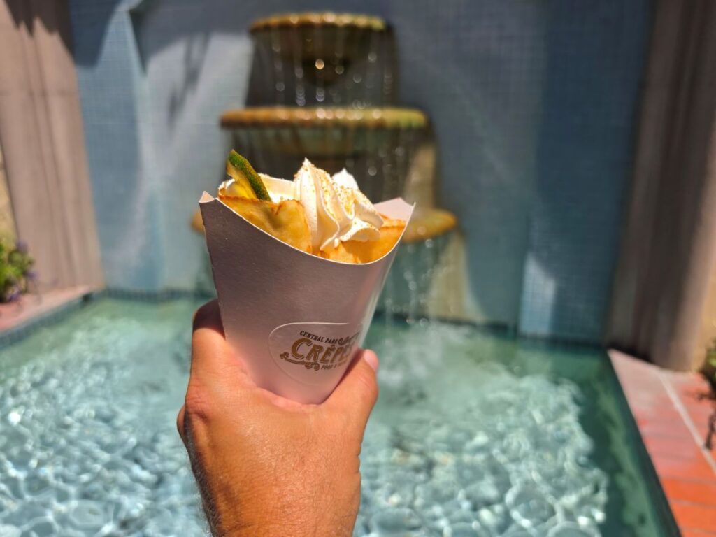A close-up view focusing on the top of a golden-brown crepe filled with whipped cream, graham cracker dust, and a fresh lime wedge, held in front of a water fountain.