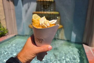 A hand holding a Key Lime crepe wrapped in a white paper cone featuring the Central Park Crêpes logo, topped with whipped cream, graham cracker crumbs, and a fresh lime slice in front of a bubbling stone fountain.