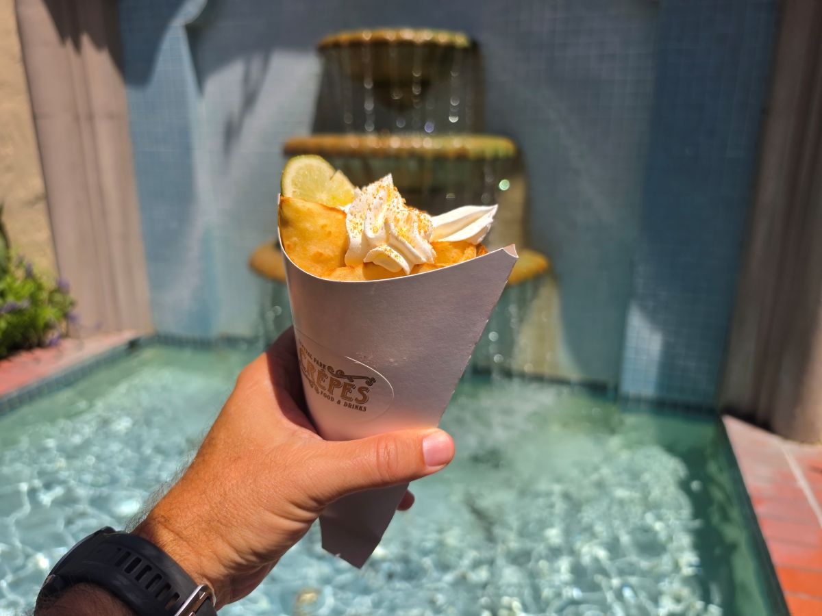 A hand holding a Key Lime crepe wrapped in a white paper cone featuring the Central Park Crêpes logo, topped with whipped cream, graham cracker crumbs, and a fresh lime slice in front of a bubbling stone fountain.