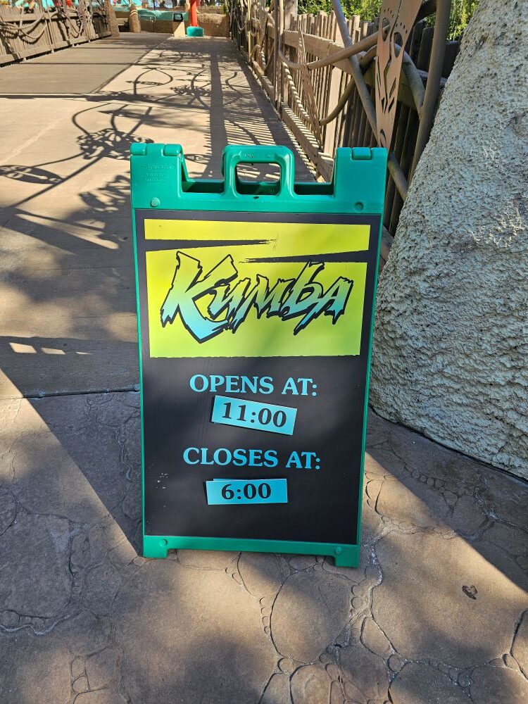 A green and black A-frame sign on a stone pathway showing the operating hours for Kumba, reading opens at 11:00 and closes at 6:00.
