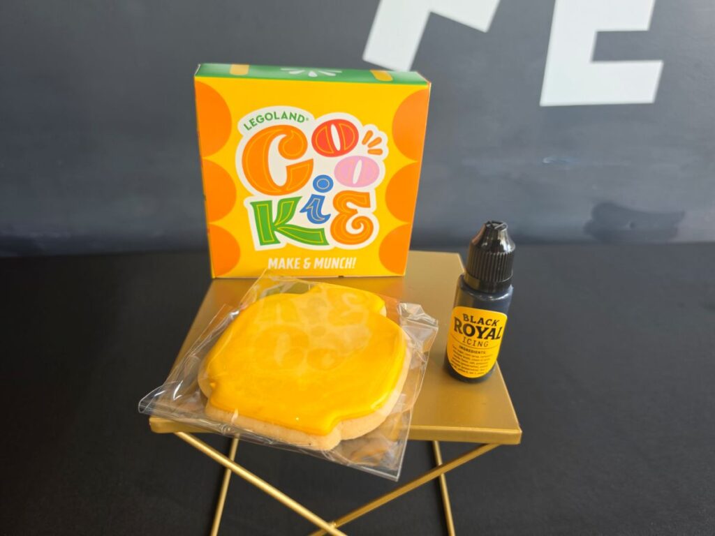 A "Decorate Your Own" Lego cookie kit setup on a gold stand, featuring a yellow-frosted minifigure head cookie in plastic wrapping, a bottle of black royal icing, and a colorful Legoland Cookie box.