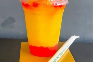 A vibrant yellow and red layered slush drink topped with red boba pearls in a clear plastic cup, resting on a small gold display stand next to a paper-wrapped compostable straw, which is one of the LEGO festival treats.