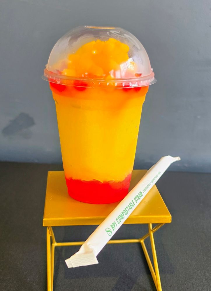 A vibrant yellow and red layered slush drink topped with red boba pearls in a clear plastic cup, resting on a small gold display stand next to a paper-wrapped compostable straw, which is one of the LEGO festival treats.
