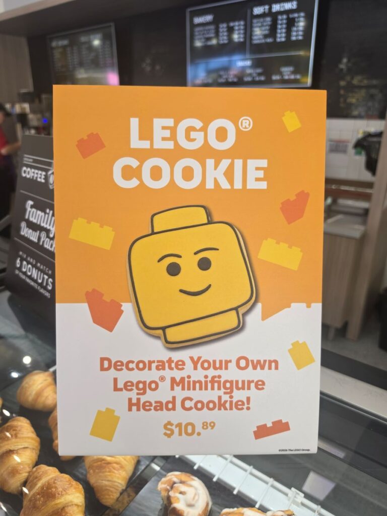 A promotional sign for "Lego Cookie" advertising a "Decorate Your Own Lego Minifigure Head Cookie!" for $10.89, featuring an illustration of a smiling yellow minifigure head cookie surrounded by Lego brick graphics for this LEGO festival treat