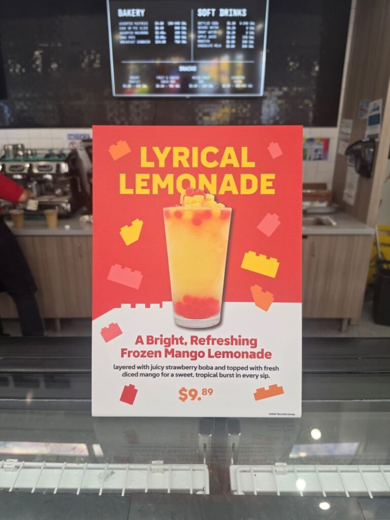 A red promotional sign advertising the "Lyrical Lemonade," described as a frozen mango lemonade layered with strawberry boba and topped with diced mango for $9.89, displayed in front of a café service counter.