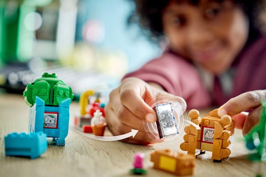 A close-up shot of a child's hands holding a clear, lit-up Lego brick with a motion graphic arrow showing it being moved from a green Lego Bulbasaur toy to a brown Lego Teddiursa toy on a wooden table.