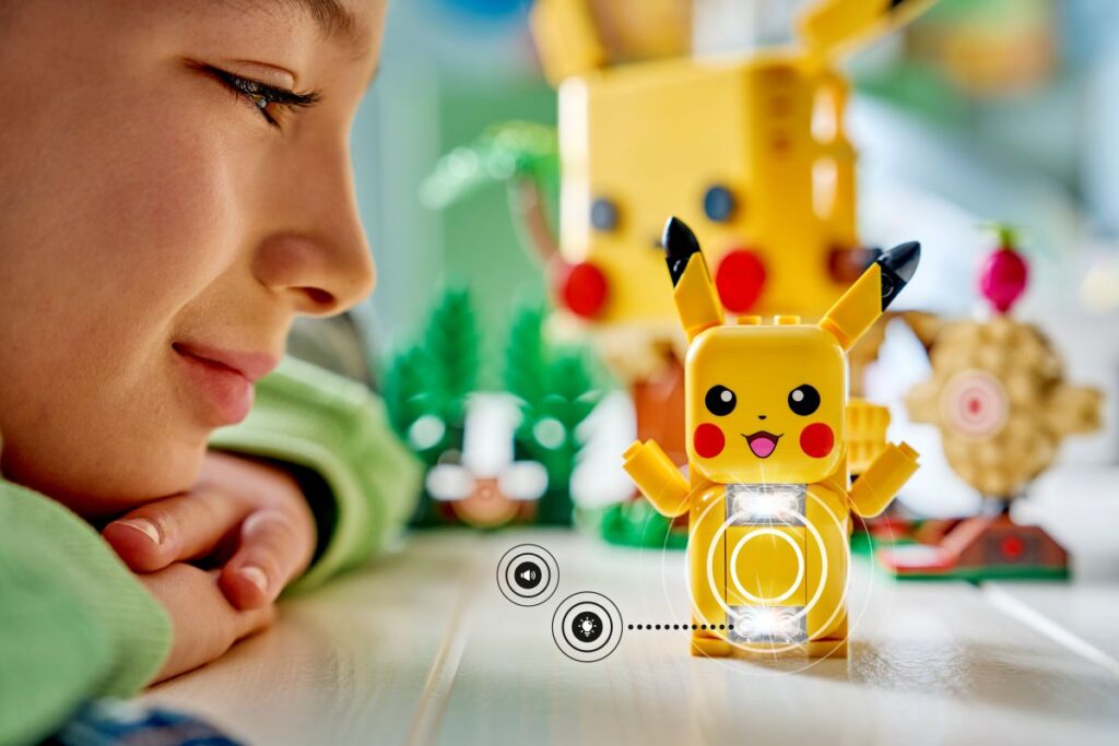 A child watches closely as a small yellow Lego Pikachu toy lights up from its belly, with circular graphic rings and small icons indicating built-in light and sound features. Promotional photo for Lego Pokemon Legoland Play Zone.