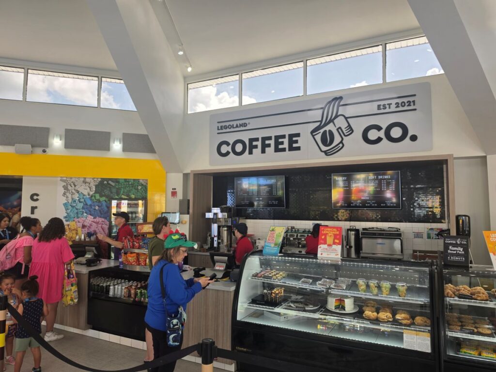 Arm & Hammer Legoland Tickets: The interior ordering counter at Legoland Coffee Co., featuring a large wall sign above the counter, glass pastry display cases filled with treats, and guests waiting in line to order.