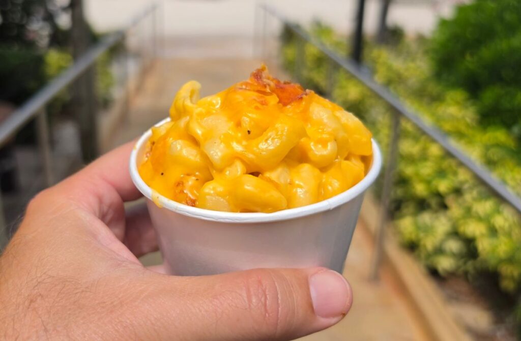 Close-up of a person holding a white paper cup filled with warm, creamy baked macaroni and cheese at Mango Joe's Beach Cafe.