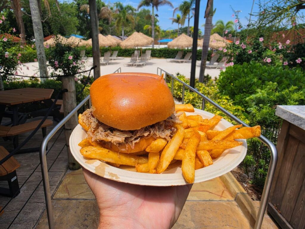 Adventure Island pulled pork sandwich and seasoned crinkle-cut fries held up against a background of outdoor theme park seating and tropical umbrellas.