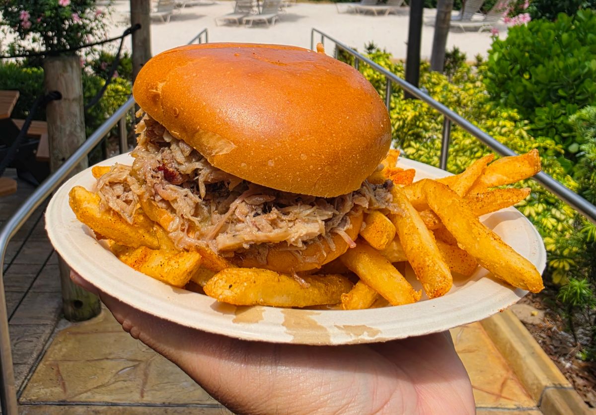 Close-up of a thick Adventure Island pulled pork sandwich on a brioche bun served with a side of seasoned french fries on a paper plate.