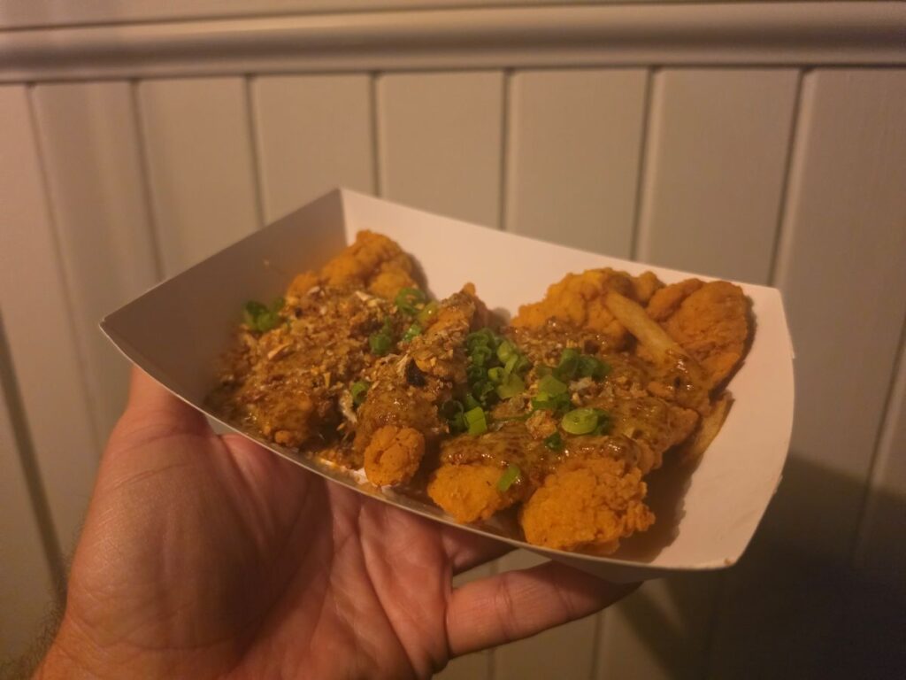 Slightly angled close-up of a hand holding crisp Maple-Mustard Chicken Strips topped with sauce and chopped green onions at Columbia Harbour House in Magic Kingdom.
