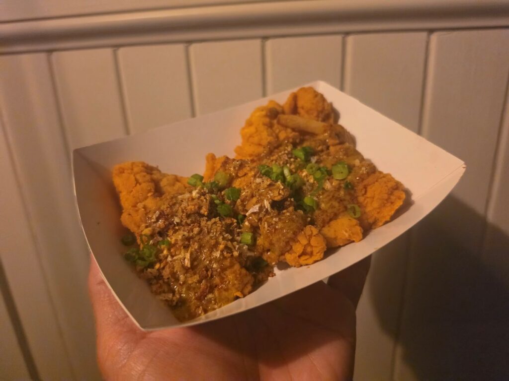Hand holding a paper boat containing Maple-Mustard Chicken Strips drizzled with sauce, crispy toppings, and green onions at Columbia Harbour House.