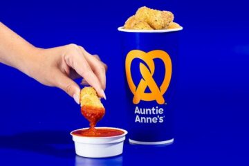 A hand dips a golden-brown pretzel nugget into a small white cup of red marinara sauce next to an Auntie Anne's cup filled with more Mozzarella Stuffed Pretzel Nuggets