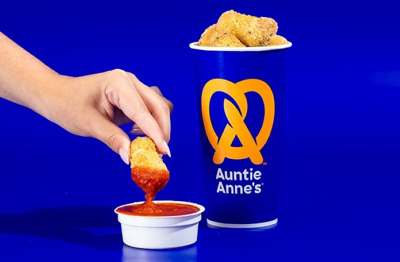 A hand dips a golden-brown pretzel nugget into a small white cup of red marinara sauce next to an Auntie Anne's cup filled with more Mozzarella Stuffed Pretzel Nuggets