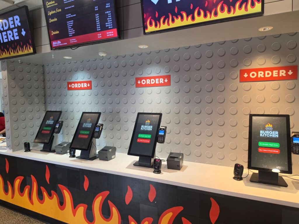 A row of four digital self-ordering kiosks inside a Burger Kitchen restaurant. The counter features an orange flame graphic, and the background wall is grey with LEGO brick details and red "ORDER" signs pointing downward.