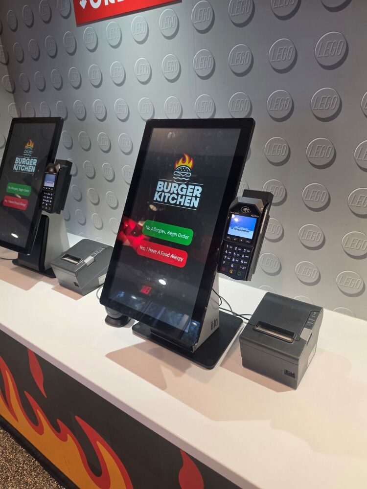 A digital self-ordering kiosk counter with a screen displaying the Burger Kitchen logo and options for "No Allergies, Begin Order" and "Yes, I Have A Food Allergy." The kiosk is positioned against a grey wall textured with LEGO brick studs.