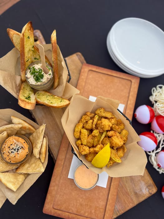 Overhead view of three appetizers served in paper-lined baskets on wooden boards. Features a dip with toasted garlic bread, a basket of fried seafood with a lemon wedge and dipping sauce, and a dish of hummus sprinkled with sesame seeds served alongside warm pita wedges.