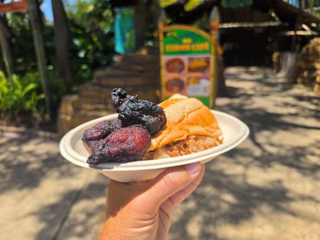 A shredded pork Pan con Lechon sandwich on Cuban bread served with fried sweet plantains on a paper plate, held up outside the Cuban Cafe at Busch Gardens Tampa Bay.