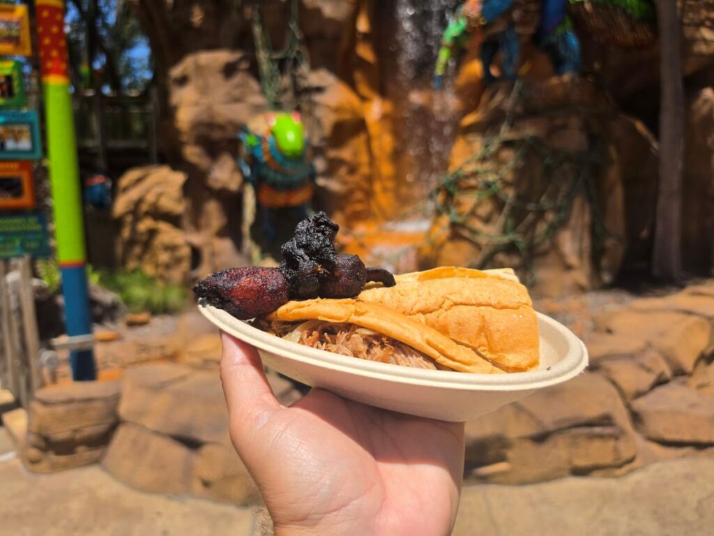A close-up view of a hand holding a Pan con Lechon Cuban pork sandwich plate topped with sweet plantains, with a rocky waterfall structure and colorful theme park decorations blurred in the background.