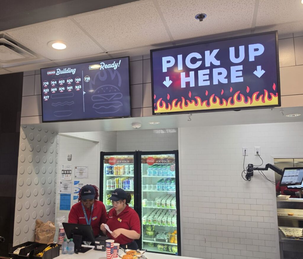 A food pickup area at a Legoland restaurant counter where two employees in red shirts and blue hats are working. Above them, digital screens display current order numbers under "Building" and "Ready!", alongside a large sign that reads "PICK UP HERE" with flame graphics for Double Cheeseburger review