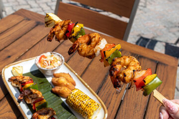 A hand lifting a grilled shrimp skewer above a full meal platter at Bob Marley – A Tribute to Freedom in honor of Jamaica Independence Day.
