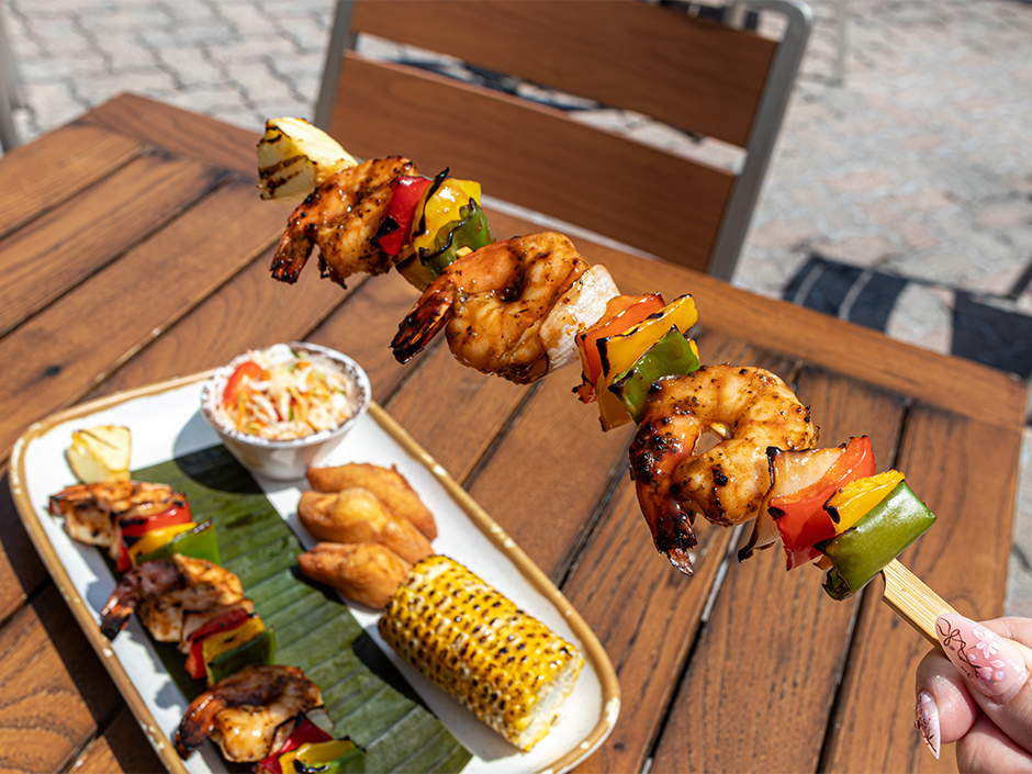 A hand lifting a grilled shrimp skewer above a full meal platter at Bob Marley – A Tribute to Freedom in honor of Jamaica Independence Day.