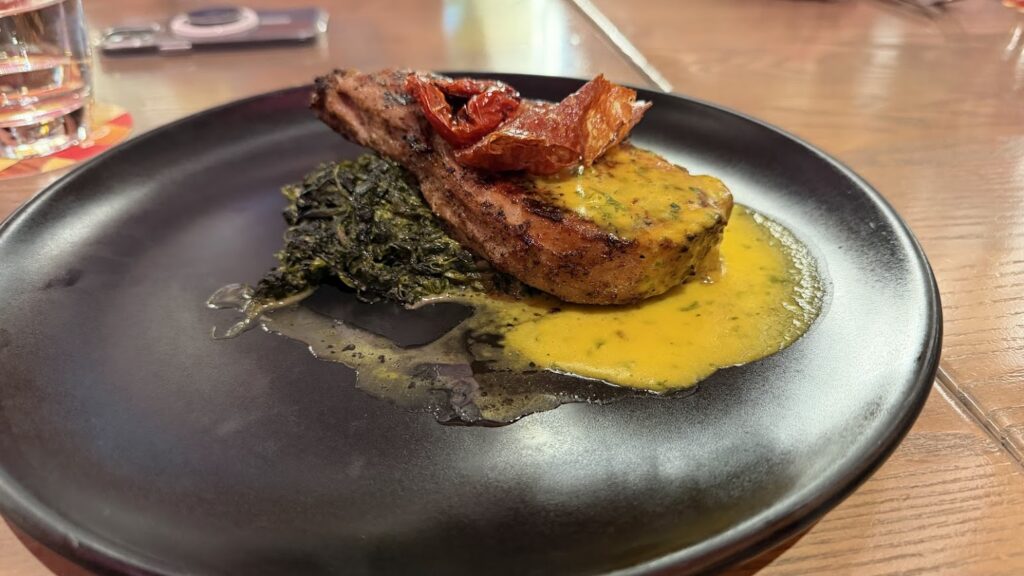 A close-up shot of a thick, seared smoked pork chop topped with crispy jamón and sun-dried tomato, served with sautéed greens and a vibrant yellow sauce on a black plate.