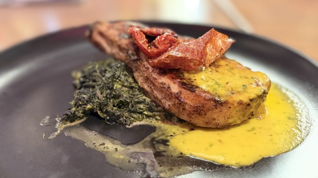 Reverse angle view of a smoked pork chop resting on a bed of dark greens, showcasing the bone-in side and a drizzle of yellow sauce on a black plate.