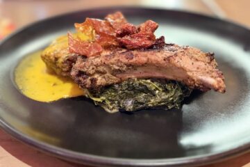 A wide-angle view of the plated smoked pork chop with crispy toppings, sautéed greens, and yellow sauce on a black plate, set on a wooden table for a Toledo Disney review.