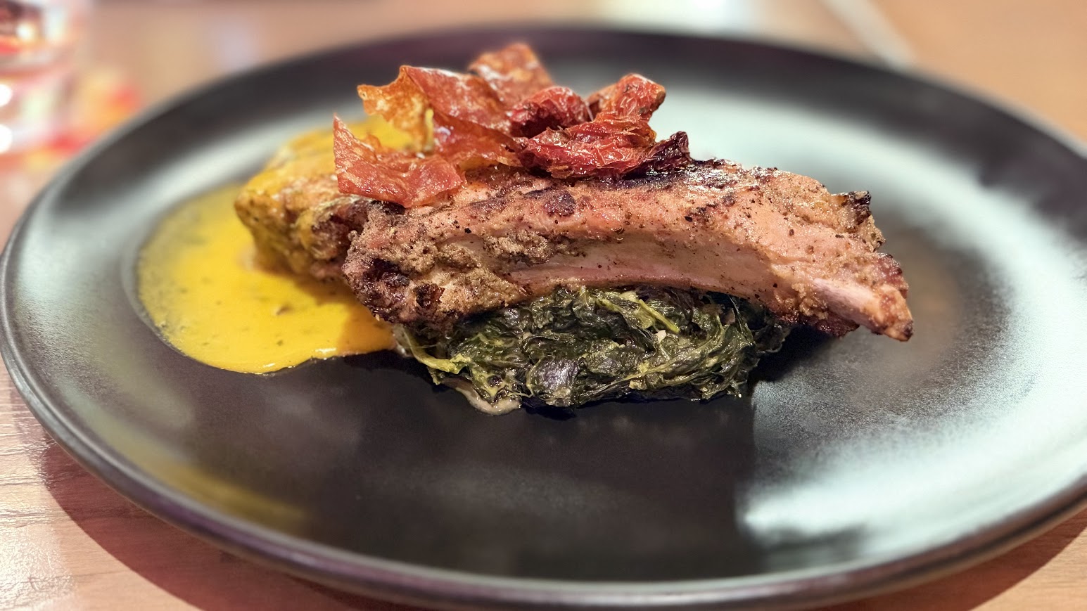 A wide-angle view of the plated smoked pork chop with crispy toppings, sautéed greens, and yellow sauce on a black plate, set on a wooden table for a Toledo Disney review.