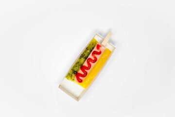 A top-down view of a Smoothie Dog from Tropical Smoothie Cafe, featuring a pink smoothie popsicle on a stick shaped like a hot dog, drizzled with red strawberry sauce and flanked by green kiwi and yellow mango purees in a small paper tray against a white background.