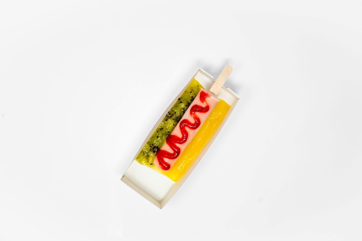 A top-down view of a Smoothie Dog from Tropical Smoothie Cafe, featuring a pink smoothie popsicle on a stick shaped like a hot dog, drizzled with red strawberry sauce and flanked by green kiwi and yellow mango purees in a small paper tray against a white background.
