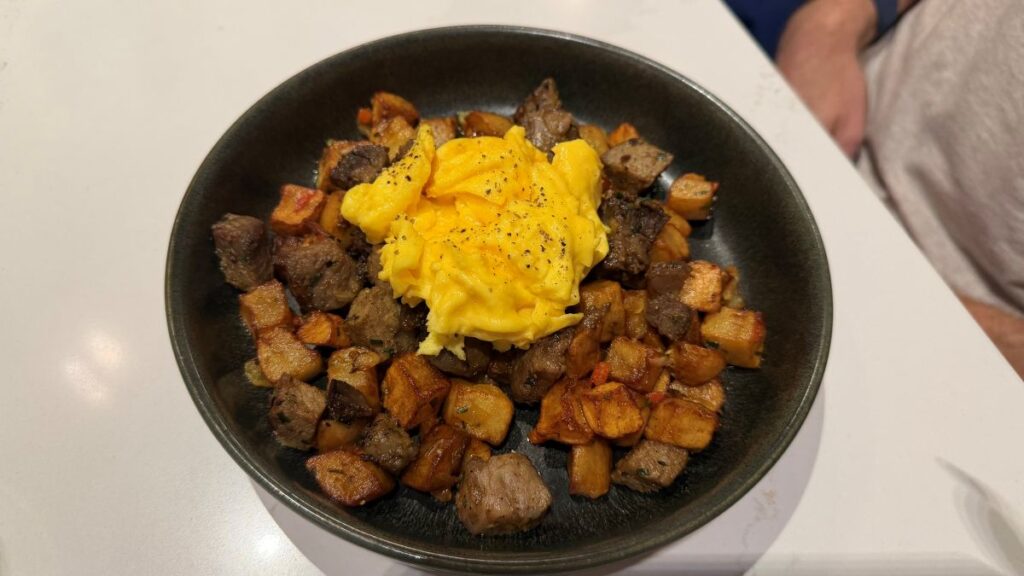 A dark ceramic bowl filled with Steakhouse 71 breakfast hash, featuring roasted cubed potatoes and tender chunks of steak, topped with a fluffy mound of scrambled eggs sprinkled with black pepper.
