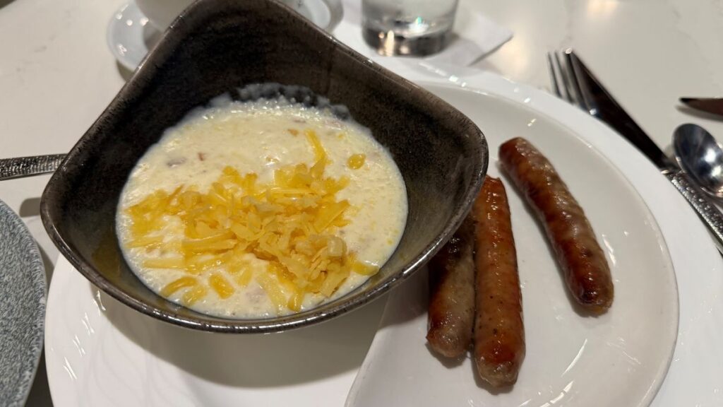 A breakfast side featuring a dark, triangular bowl of creamy, warm grits topped with shredded cheddar cheese, served alongside three browned pork sausage links on a white plate.