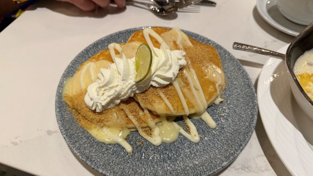 A stack of Key Lime Pie pancakes on a speckled blue plate, drizzled with key lime curd and sweet cream glaze, dusted with graham cracker crumbs, and garnished with whipped cream and a fresh lime slice.