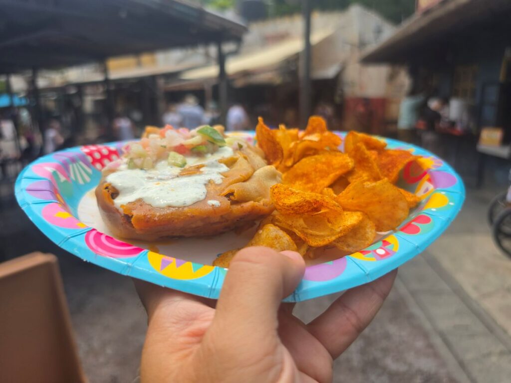 A hand holding a colorful paper plate with a Tanzanian Meat Pie topped with white sauce and fresh salsa, served with a side of seasoned chips.