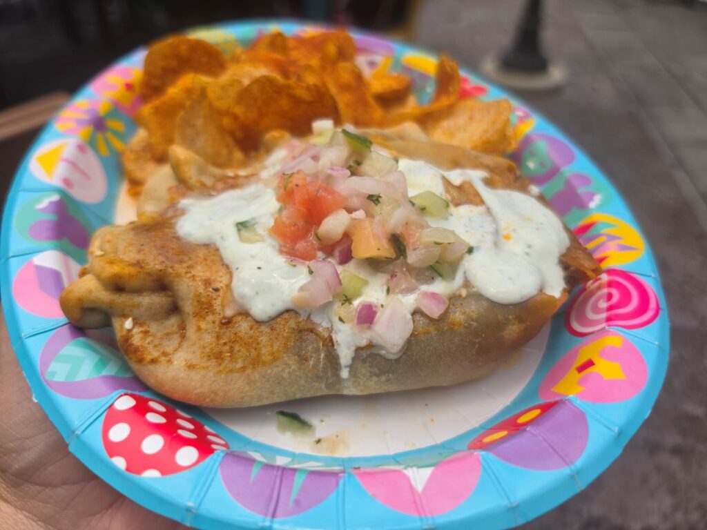 A detailed top-down view of a Tanzanian Meat Pie on a themed Disney Parks paper plate, highlighting the fresh salsa and creamy white sauce topping.