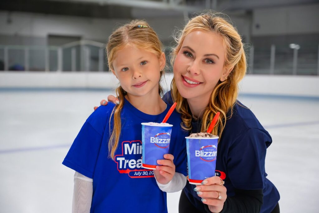 Tara Lipinski and a young girl stand on an ice rink, both smiling and holding Dairy Queen Blizzard cups with red spoons while wearing matching blue shirts for Miracle Treat Day promoton.