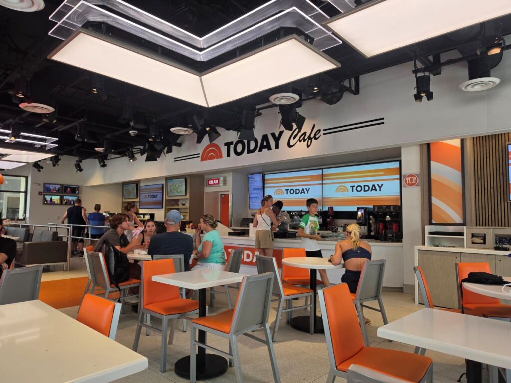 Indoor dining area of TODAY Cafe at Universal Studios Florida featuring orange chairs, white tables, and guests ordering food.