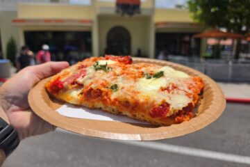 Hand holding a thick-crust Tomato Mozzarella Flatbread slice with melted cheese and fresh herbs outside TODAY Cafe at Universal Studios Florida.