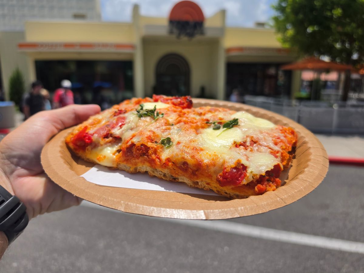 Hand holding a thick-crust Tomato Mozzarella Flatbread slice with melted cheese and fresh herbs outside TODAY Cafe at Universal Studios Florida.