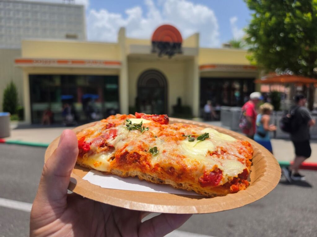 Person holding a paper plate with a Tomato Mozzarella Flatbread in front of the blurred exterior of TODAY Cafe.