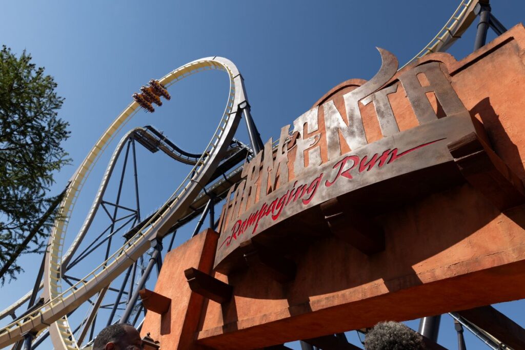A dramatic, low-angle view of a large, rustic orange entrance archway featuring the metallic sign "TORMENTA Rampaging Run." In the background, Tormenta: Rampaging Run roller coaster train climbs a massive loop on the cream and black steel track.